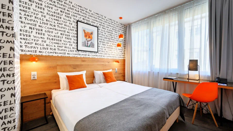 Double bed, orange accents and patterned wallpaper in a Roombach standard room.