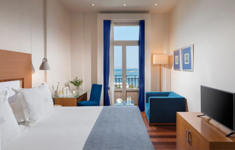 Bed, blue seating and a sea-facing window in an Executive Room – Sea View at GDM Megaron.