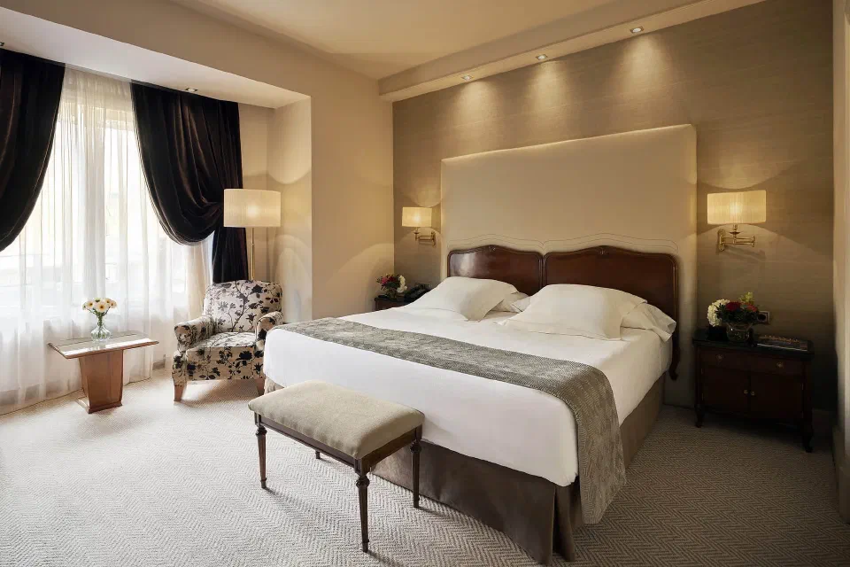 A white bed, floral armchair and dark curtains in an Executive Room at Wellington Hotel & Spa Madrid.