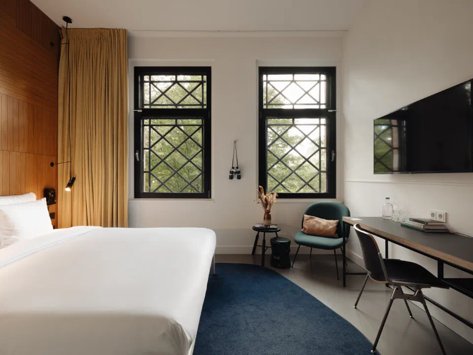 A Double Room at Conscious Hotel Westerpark, with tall patterned windows, a bed and a desk.