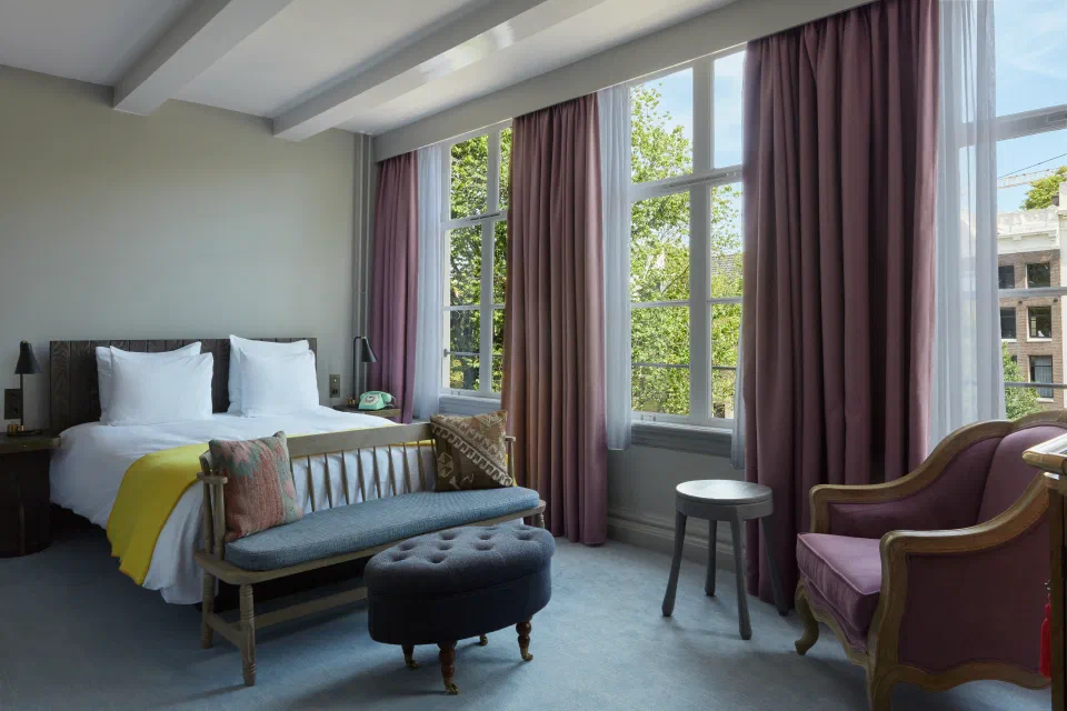 A Pulitzer Amsterdam bedroom with tall windows, pale green walls and a bench at the foot of the bed.