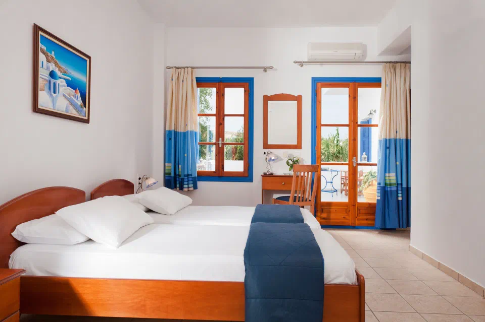 Twin beds in a white room with a blue-framed window and balcony doorway at Hotel Eleni.
