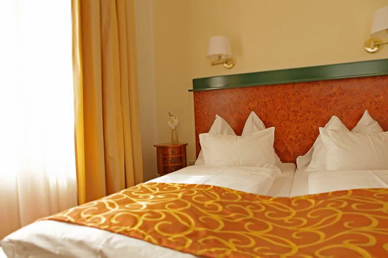 A neatly made double bed at Hotel Domizil, with warm wood and an orange patterned runner.