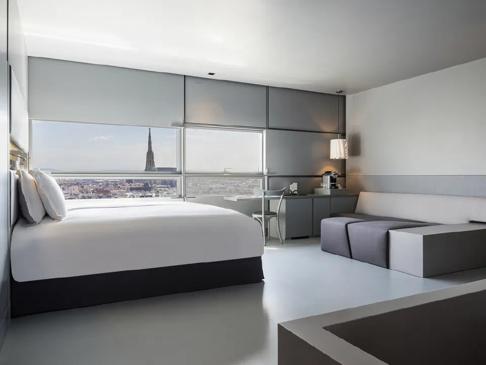 A calm, light-toned SO/ Vienna guest room with floor-to-ceiling windows and Vienna rooftops beyond.