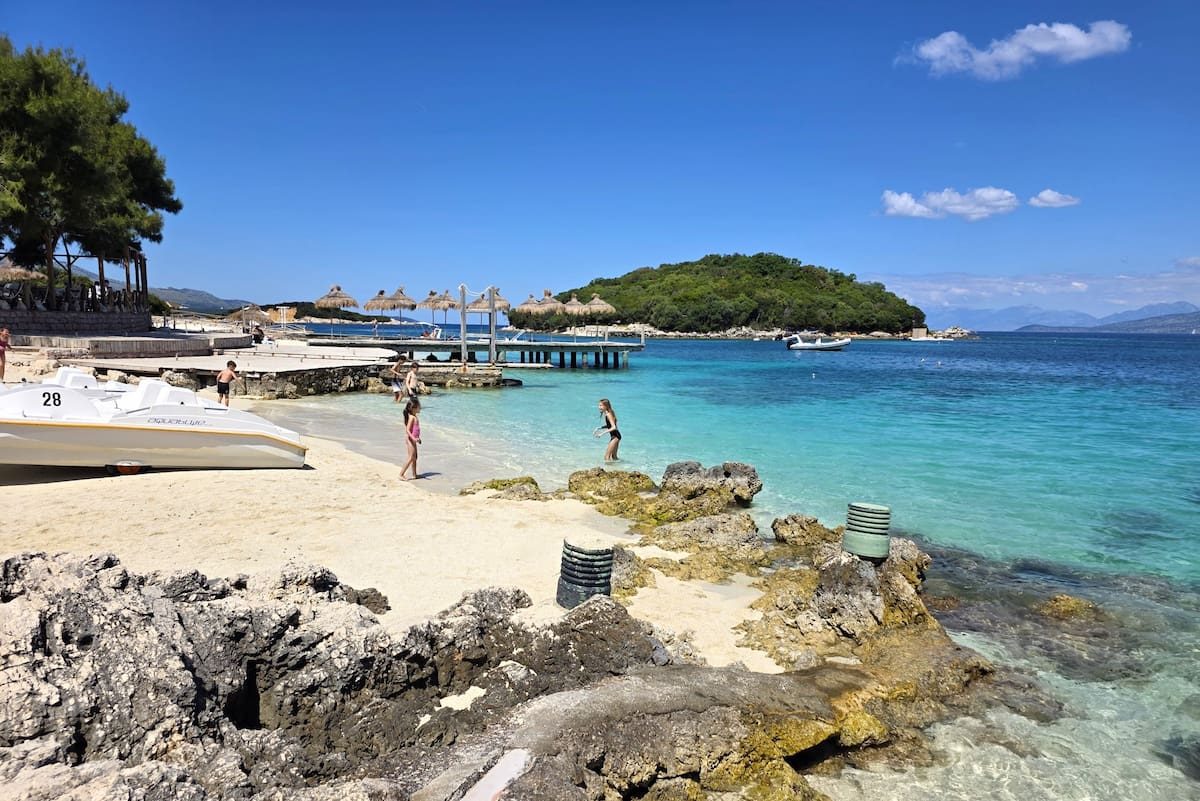White sand and clear water in a Ksamil cove, with straw parasols and an islet opposite