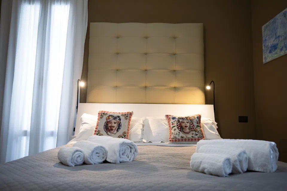 White pillows and patterned cushions beneath a padded headboard, beside a bright window in a Deluxe room at Villa Calipso.