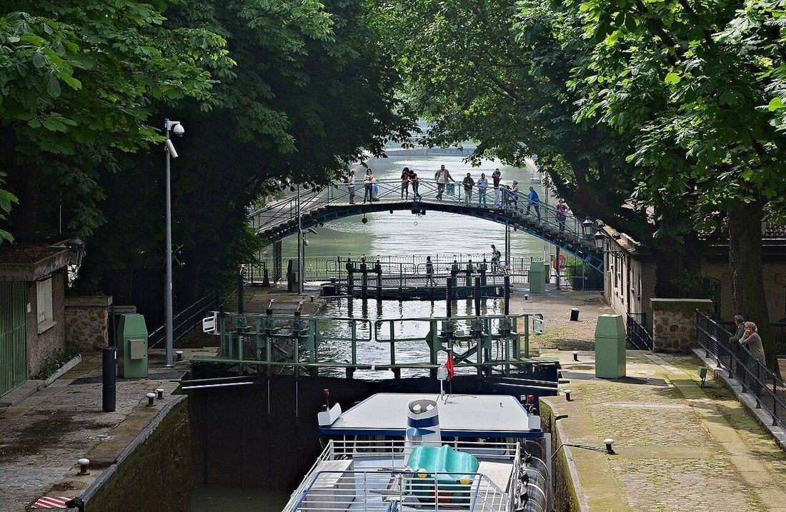 A boat at the Récollets locks on Canal Saint-Martin beneath an iron footbridge and leafy trees.