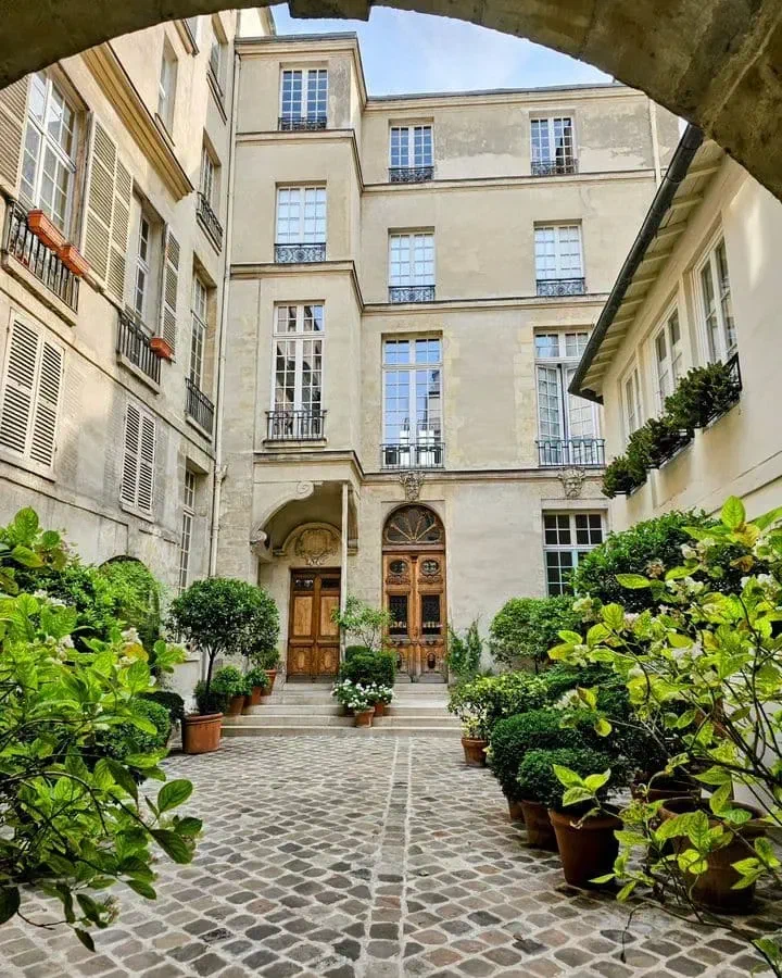 Potted plants line a stone courtyard beneath an archway, with tall windows and wooden doors.
