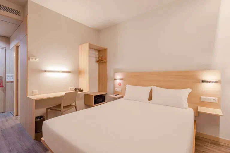 A simple double room with white bedding, a pale wood headboard and a desk at Moov Hotel Porto Centro.