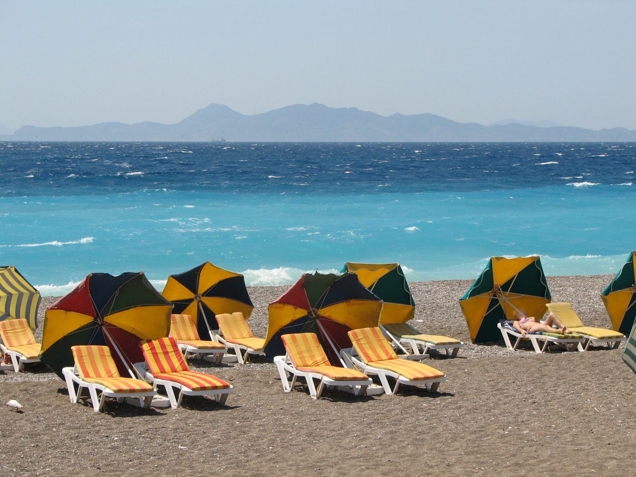Striped loungers and umbrellas on a pebbly beach beside turquoise water on Rhodes’s west coast.
