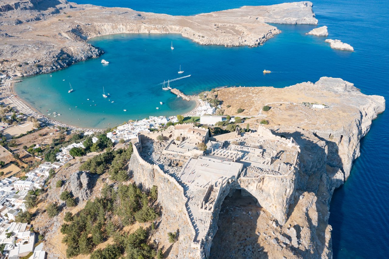 Aerial view of Lindos Acropolis above white houses and the turquoise curve of Lindos beach.