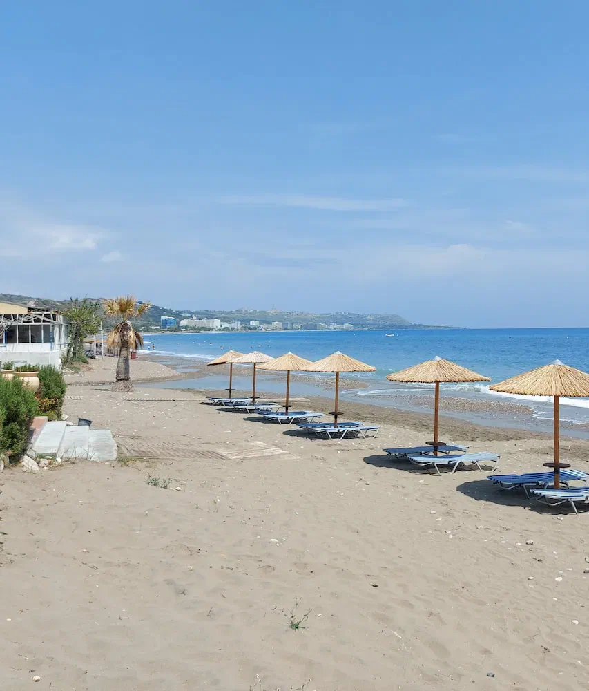 Faliraki’s sandy beach with straw parasols, the blue bay and buildings in the distance.