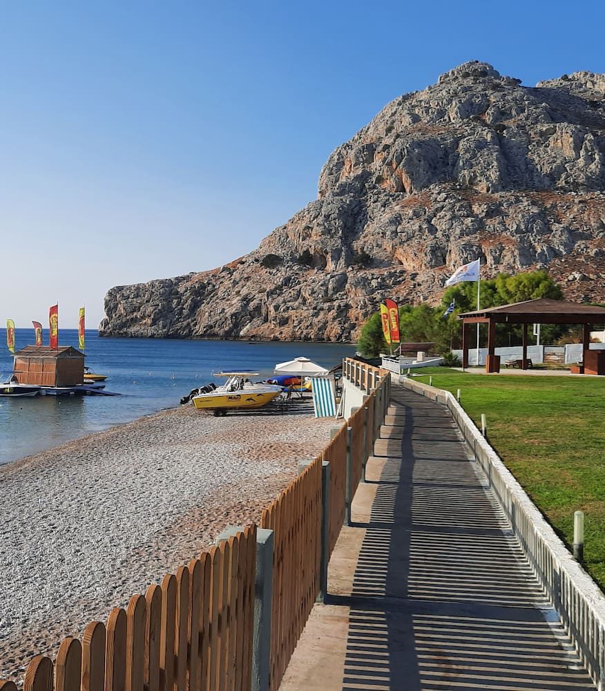 A boardwalk beside the shingle beach at Kolymbia, with boats and a rocky headland beyond.