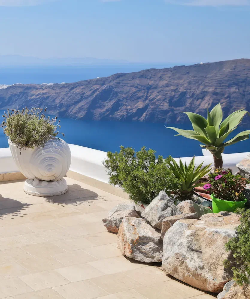 A sunlit terrace with a white plant pot overlooks the sea and caldera cliffs in Imerovigli.