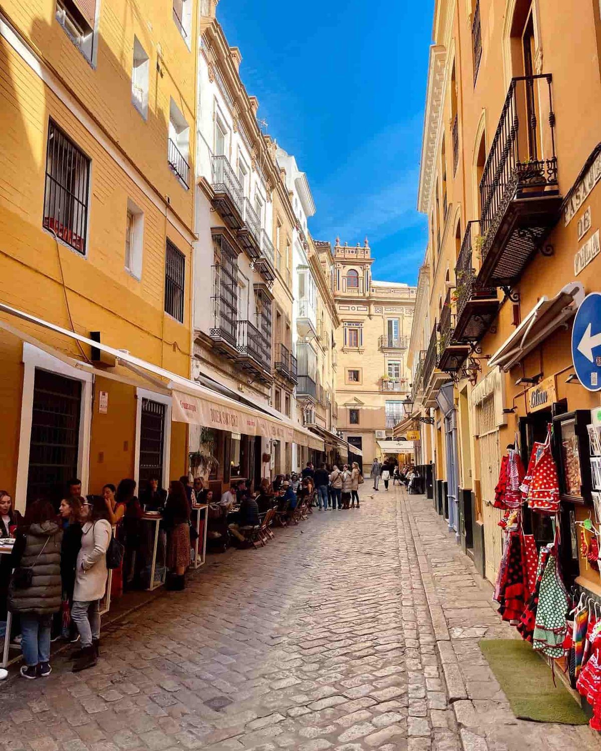 A paved lane in Seville’s Santa Cruz quarter, with bodega tables and flamenco dresses on display.